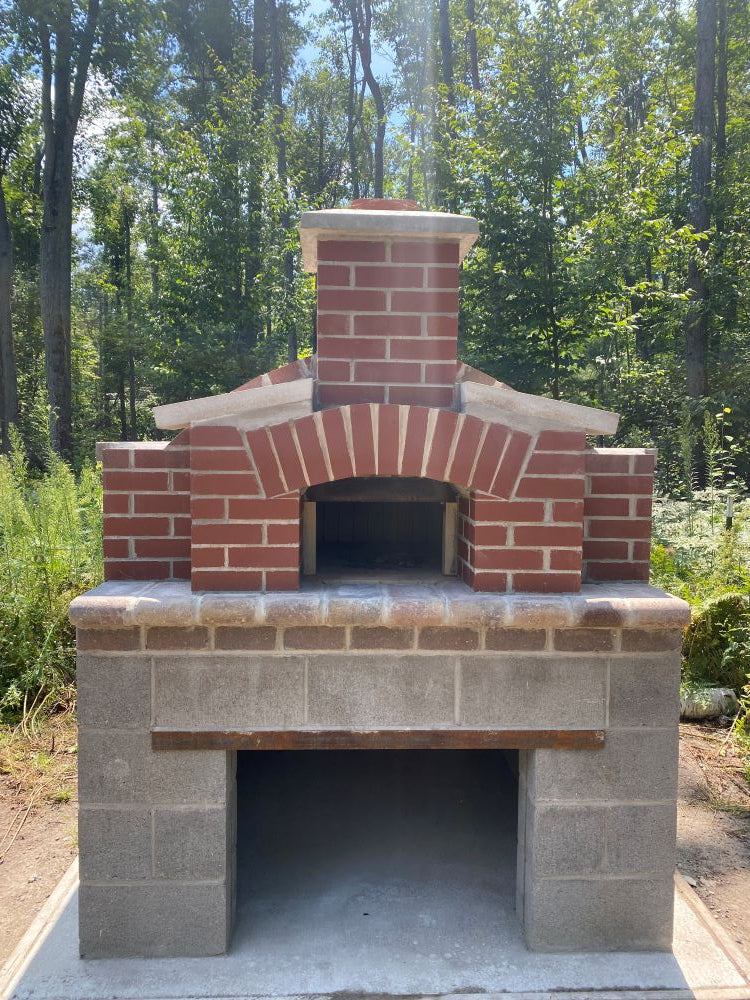A brick and stone outdoor pizza oven set against a backdrop of dense green trees. The oven features a red brick chimney and arched opening.